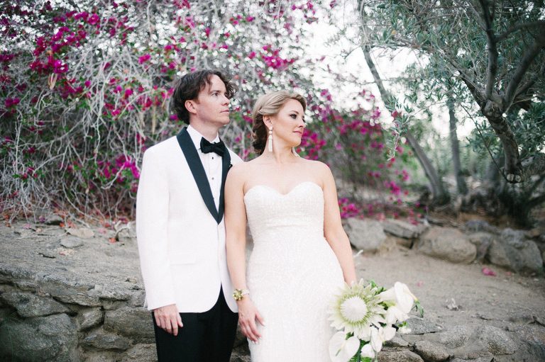 Married couple man with white tux and black bowtie woman in Wtoo sparkling wedding gown, beaded drop earrings, protea bouquet, bougainvillea in background