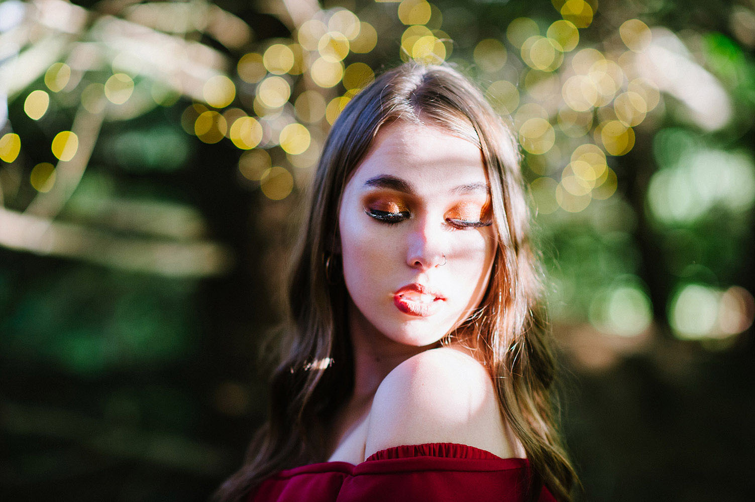 Girl with long brown hair and off the shoulder dark red top looking down with umber eye shadow and lipstick and bokeh lights background