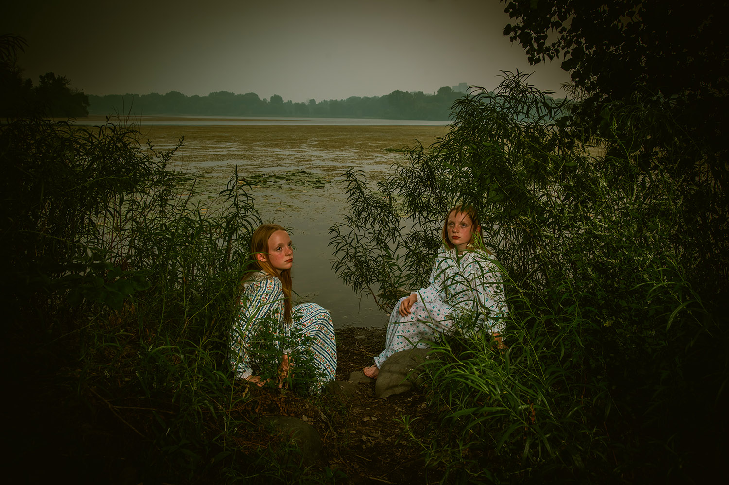 Twin girls sit in the weeds next to a lake in nightgowns with an eerie vibe.