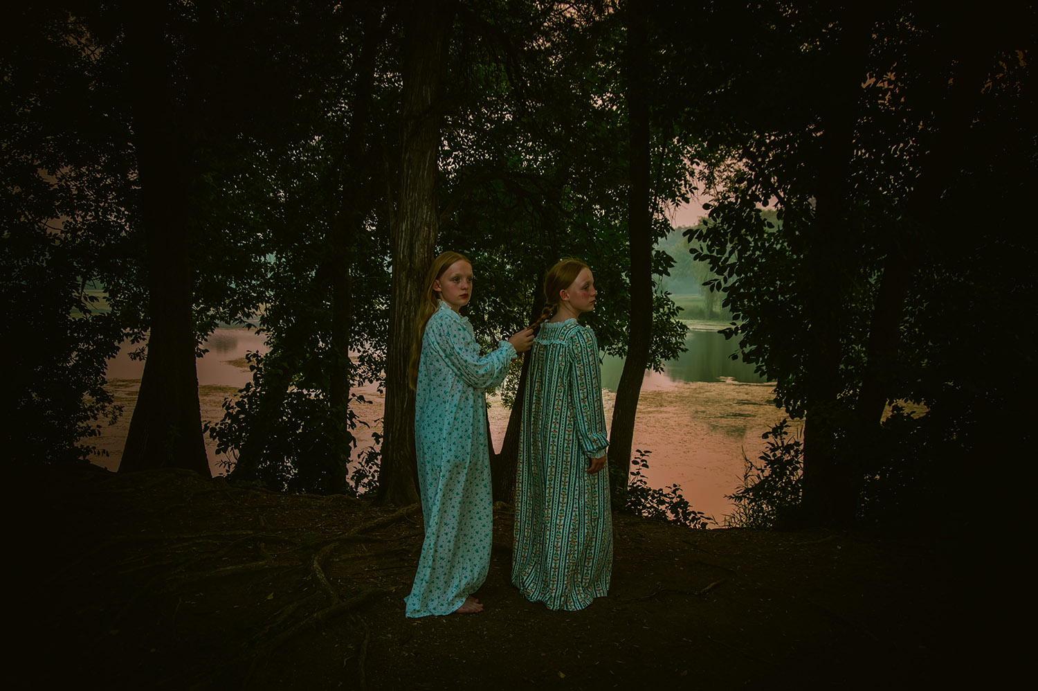 Twins stand in a woods in nightgowns by a lake with an eerie vibe