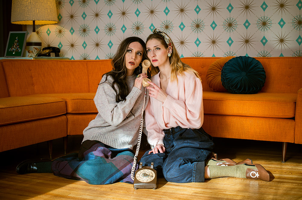 two girls sit on the floor in front of an orange couch listening to an old telephone