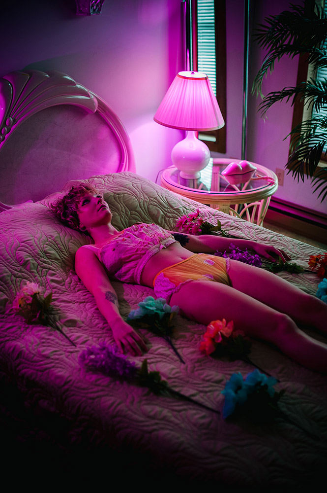 a girl lies on a bed in her underwear under pink light surrounded by fake flowers