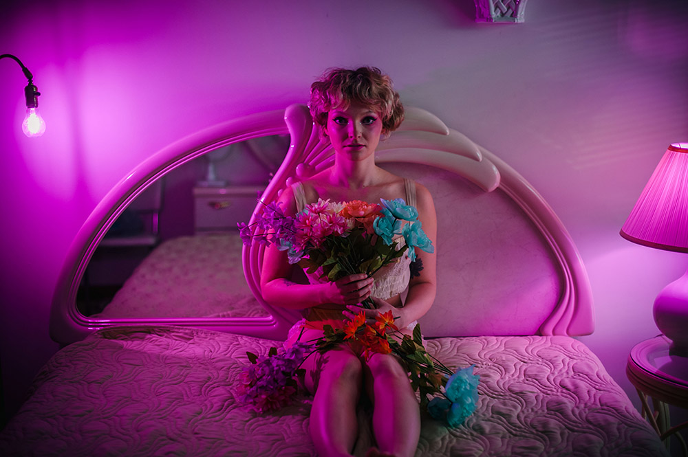 a girl sits on a bed under pink light holding fake flowers