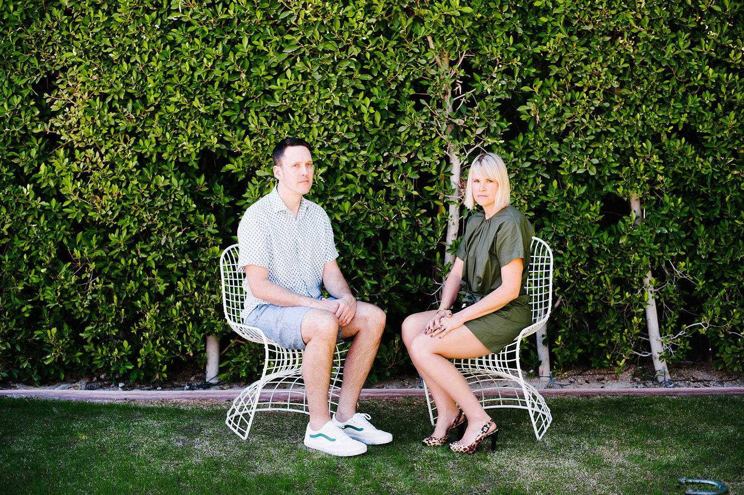woman in green jumper with leopard print heels and guy in white shirt with blue dots in blue shorts in white wire chairs in front of green bush wall