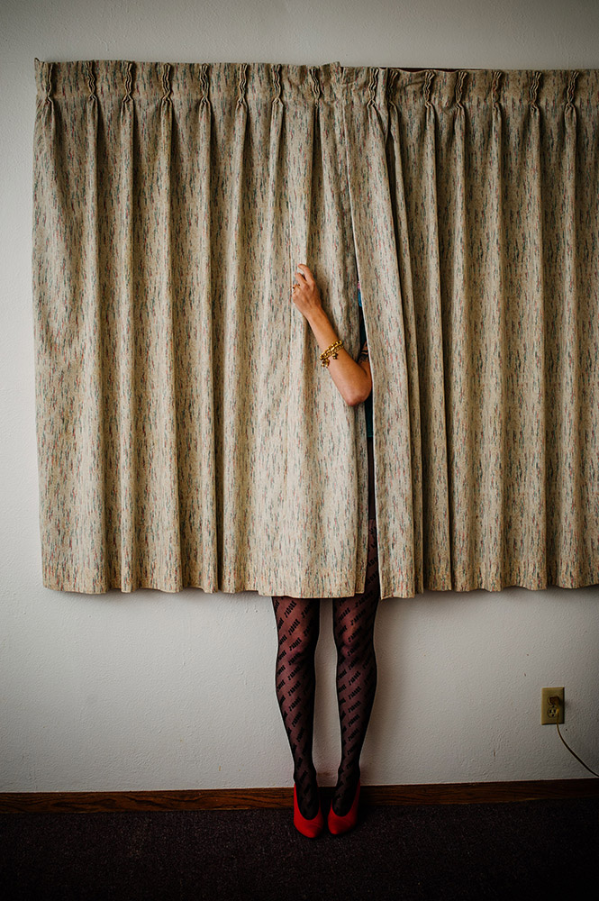 a womans arm sticks out from behind beige curtains and she is wearing black tights and red shoes
