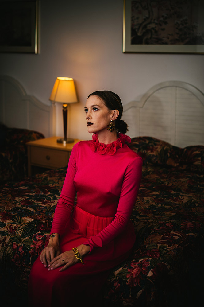 a woman sits on an old motel bed wearing a vintage pink dress staring off into space