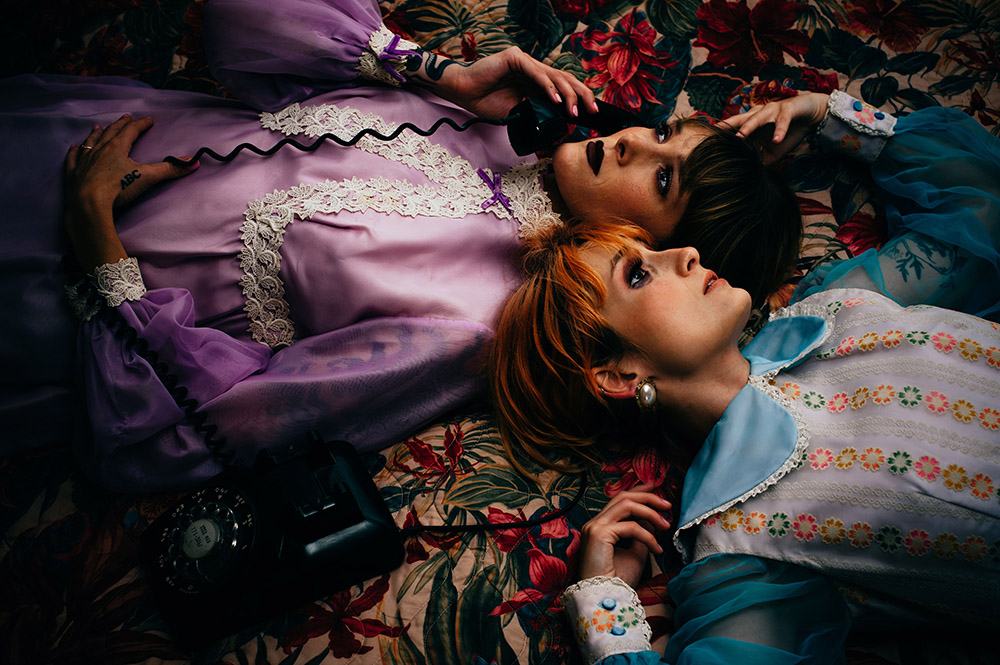 two girls lying on an old hotel bed one in a blue dress one in a pink dress and holding a vintage phone