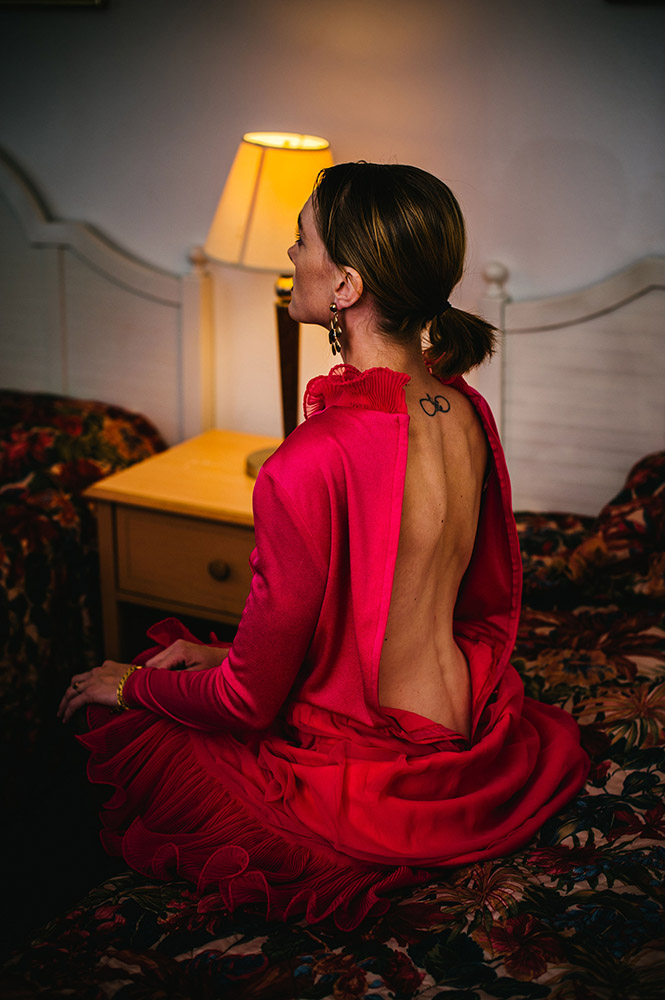 a woman sits on an old motel bed facing away from us wearing a vintage pink dress with the back unzipped