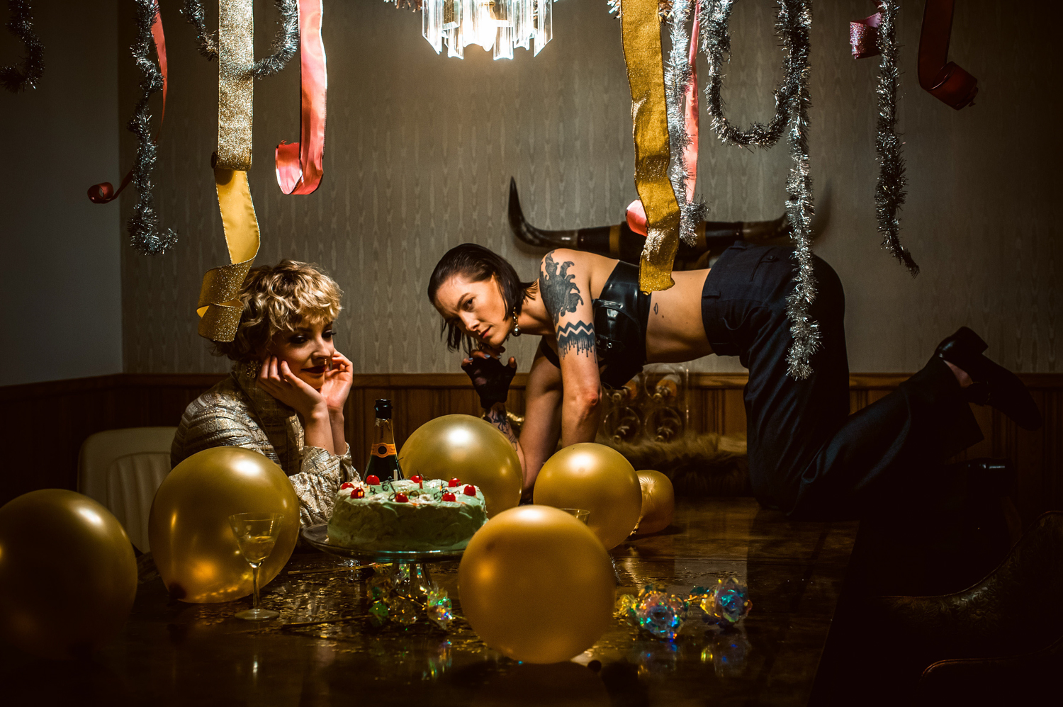 two white women at a table one starring at a cake and one on the table in bondage bra at Grandpas Pool House