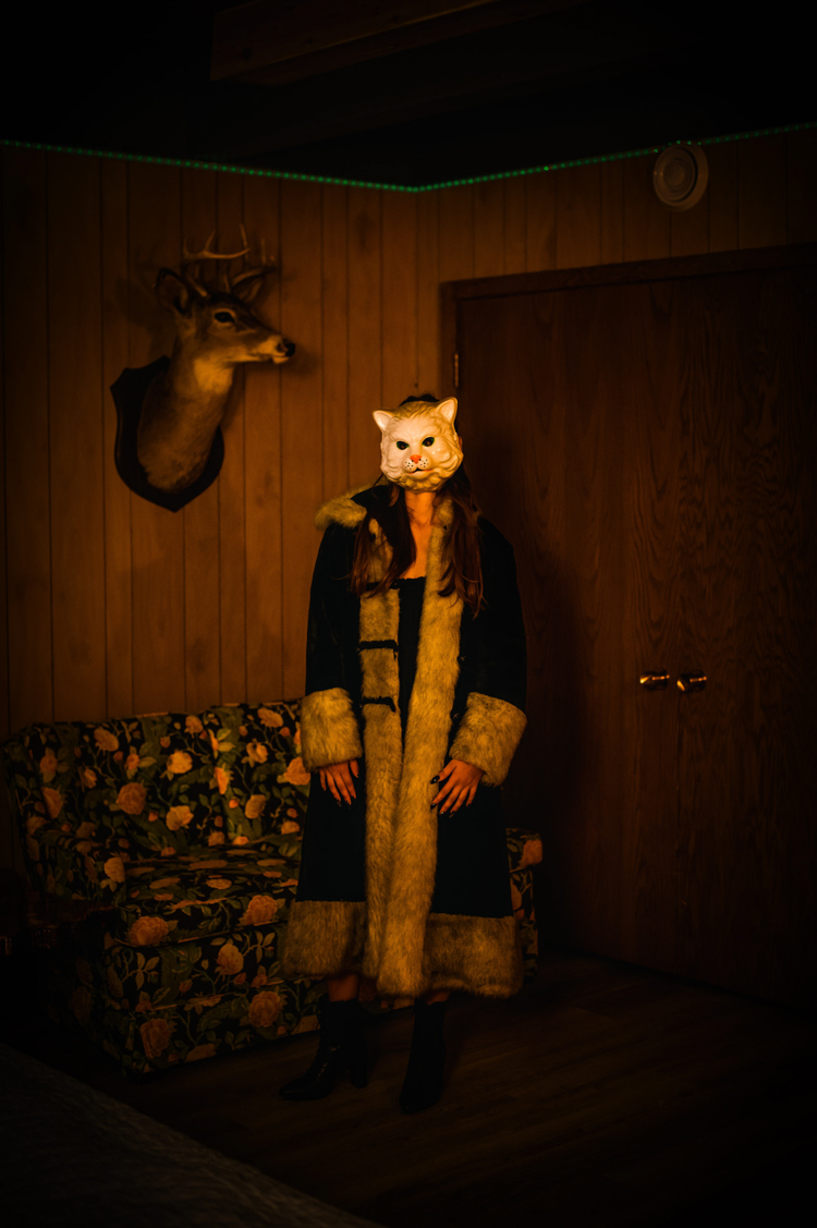 a person in a white cat mask an a long Narnia style coat standing next to a mounted deer head at Grandpas pool house