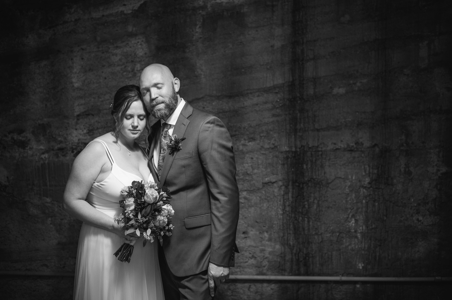 a black and white photo of a married couple closing their eyes in front of a cement wall